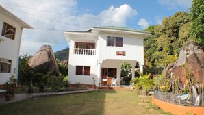 A two-story white house with a balcony, surrounded by lush greenery and large rocks.