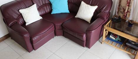 A maroon leather sectional sofa with two white pillows and one blue pillow, positioned on a tiled floor with a small wooden table and a vase of dried flowers nearby.
