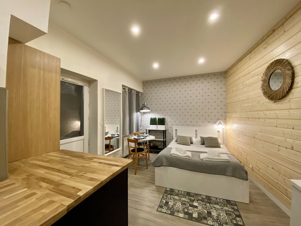 A compact bedroom with a wooden headboard, a bedside table, a bed with pillows, a small desk with a chair, and a mirror on the wall.