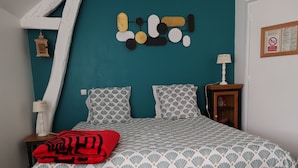 A bedroom with a teal accent wall, a patterned bedspread, two matching pillows, a red throw blanket, a nightstand with a lamp, and a framed sign on the wall.