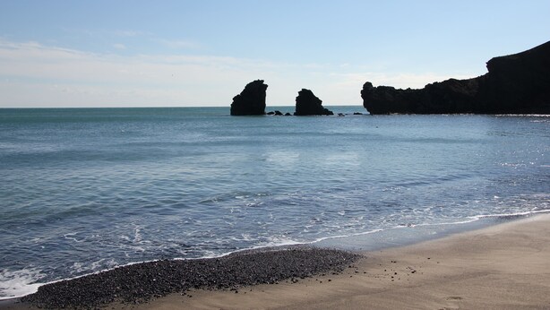 Una playa con aguas tranquilas, orilla pedregosa y formaciones rocosas al fondo.
