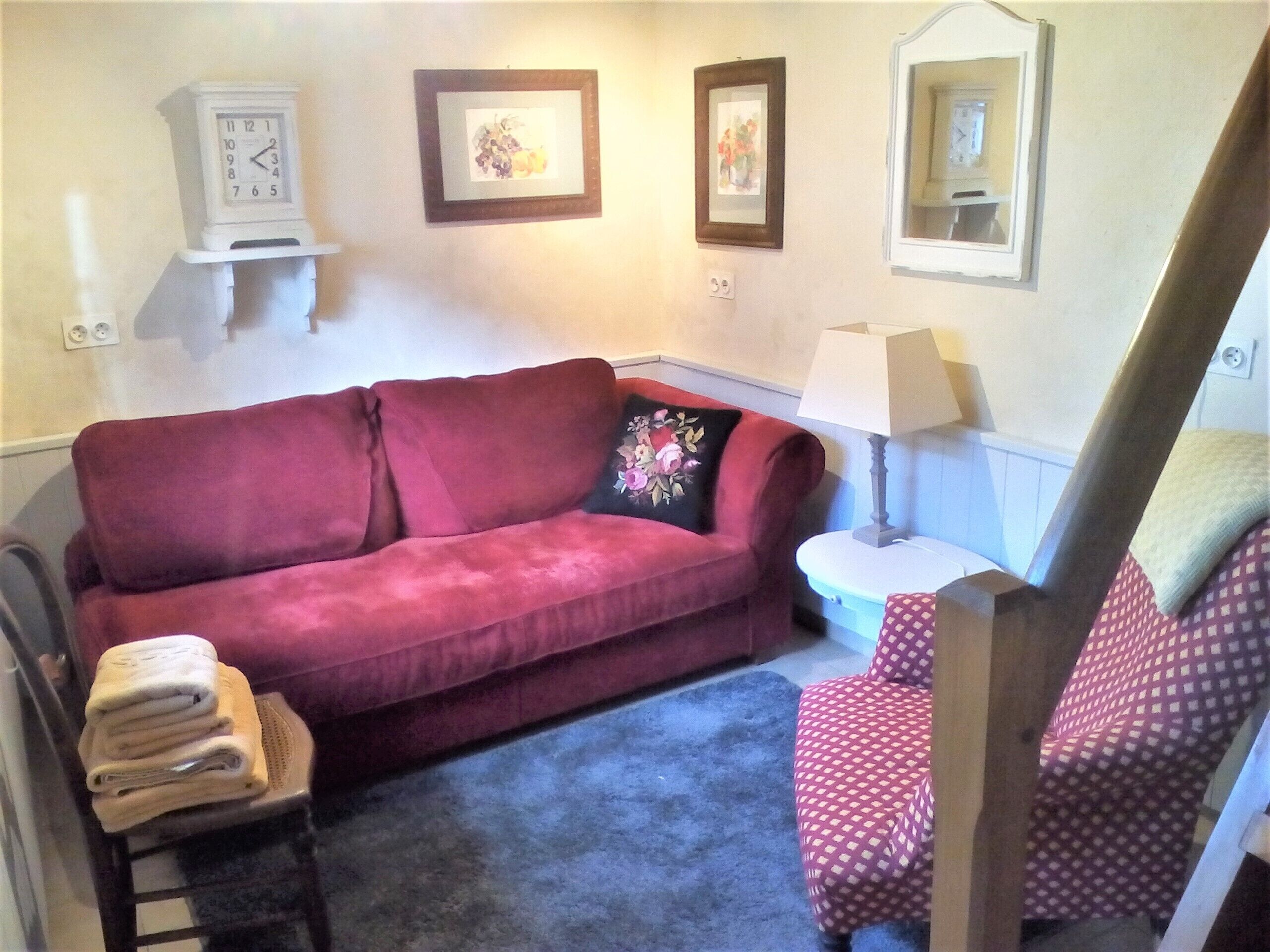 A living room with a red sofa, a patterned armchair, a small table with a lamp, and wall-mounted clocks.