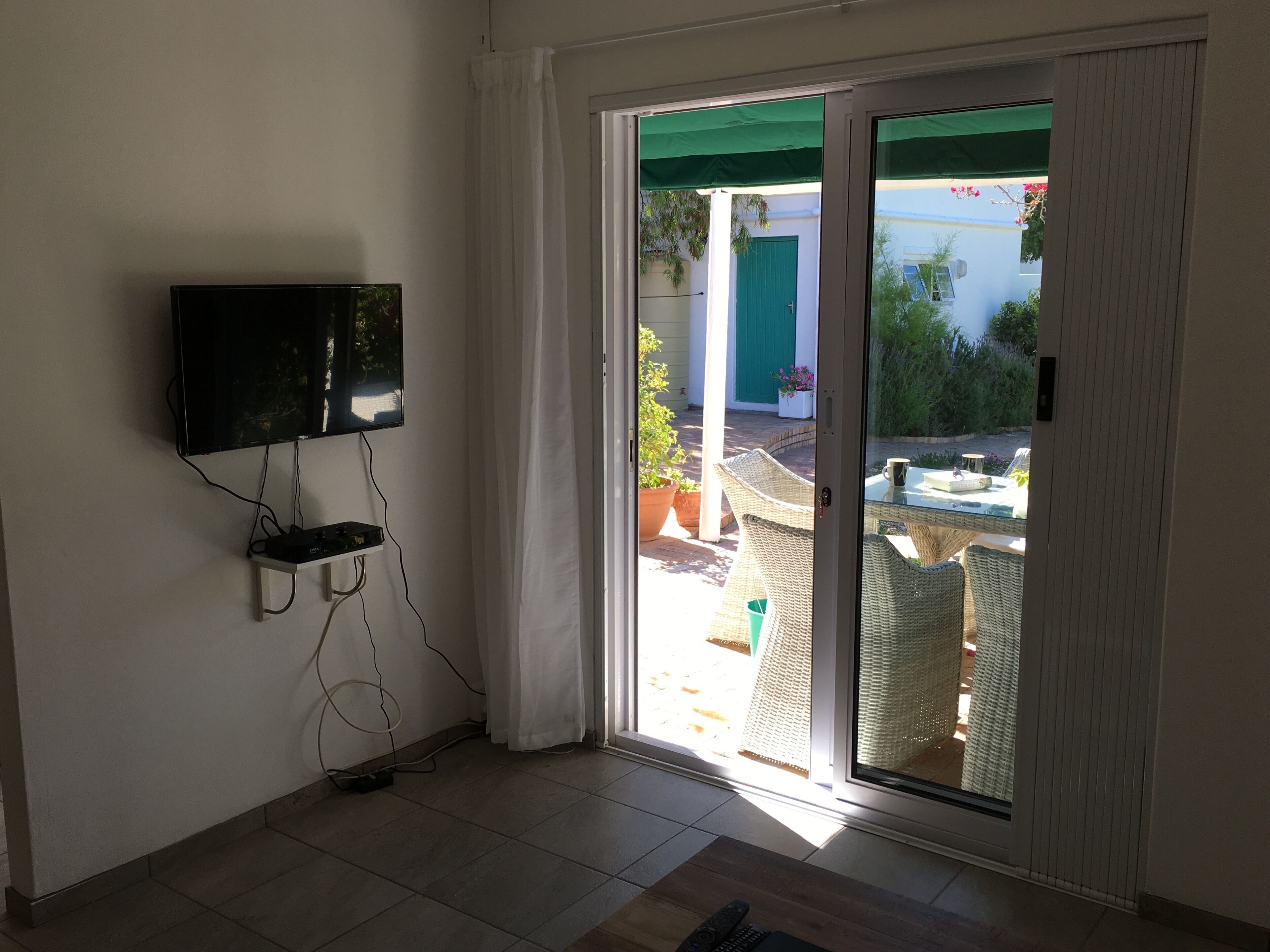 Une pièce dotée d’un téléviseur fixé au mur, d’une porte-fenêtre coulissante donnant sur un patio extérieur aménagé avec des meubles en osier et des plantes en pot.