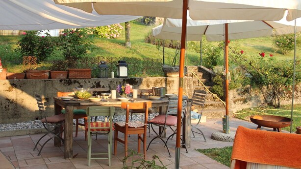 Un patio al aire libre con una mesa y sillas bajo un paraguas grande, rodeado de follaje y un muro de piedra.