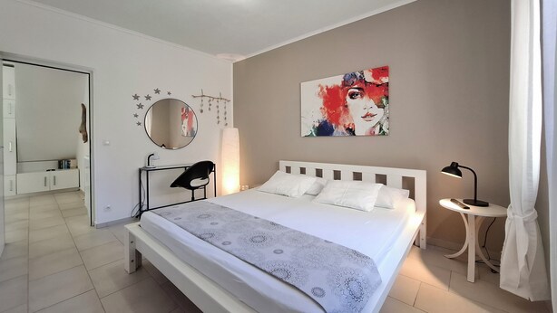 Een moderne slaapkamer met een groot bed, een nachtkastje met een lamp, een spiegel en een kunstwerk aan de muur.