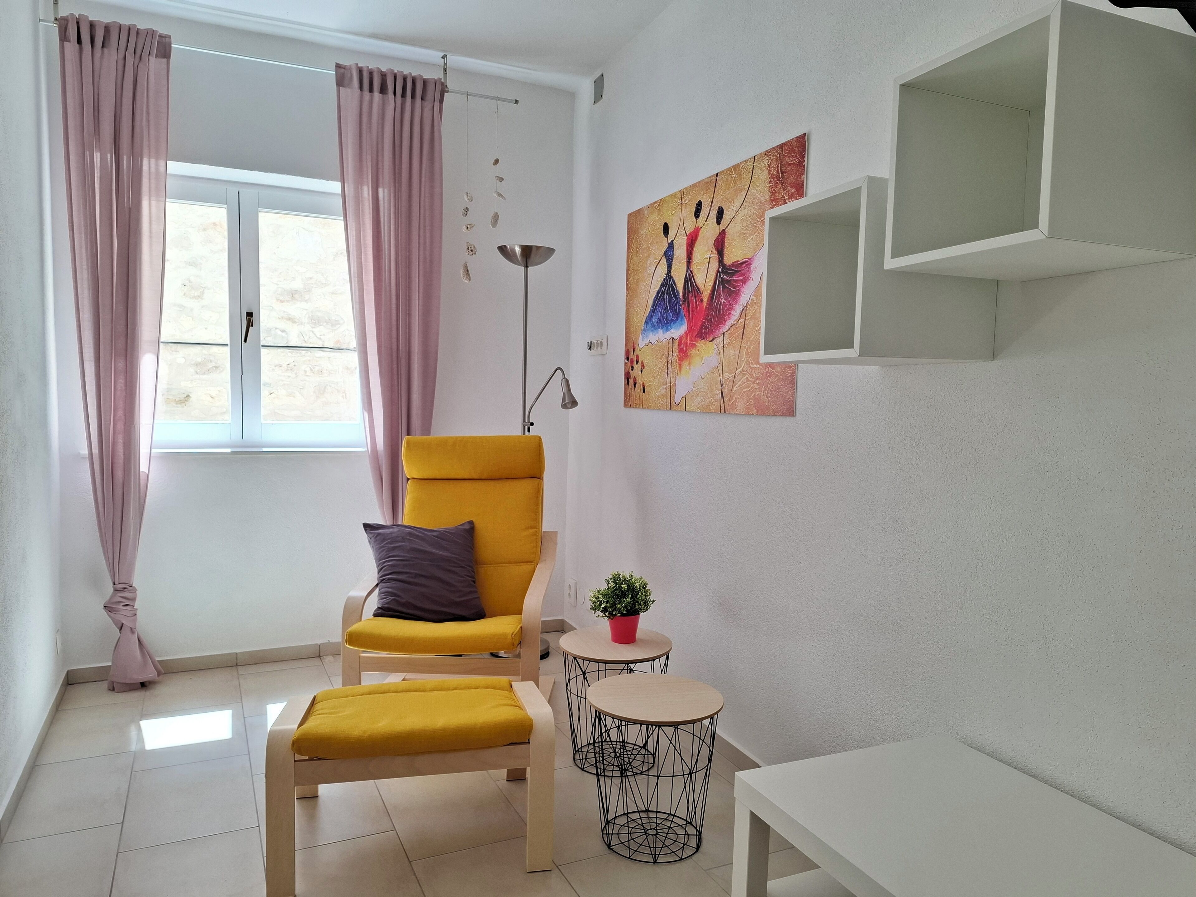 A modern living room with a yellow armchair, a small round table with a plant, and a painting on the wall.