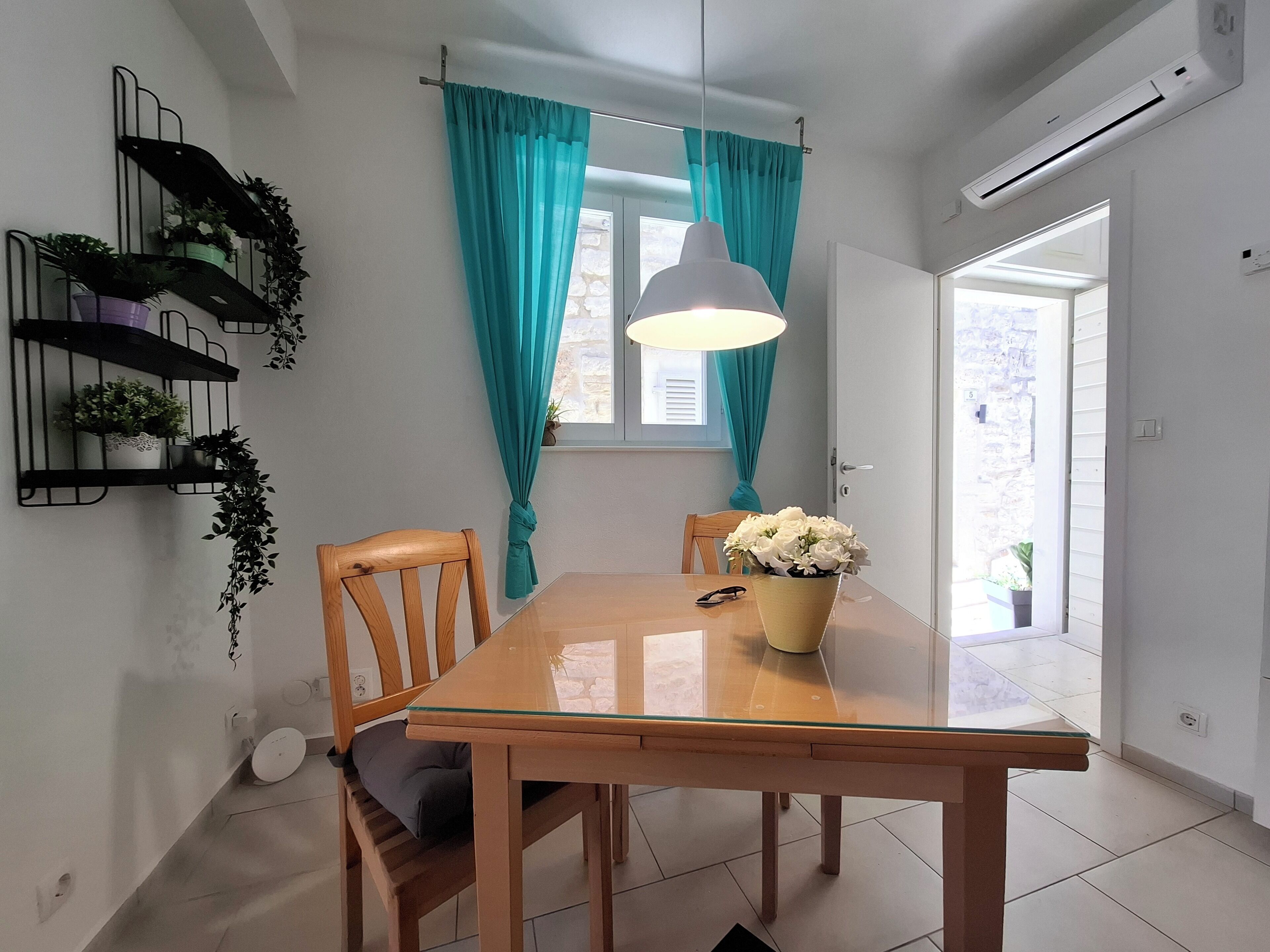Un coin repas avec une table en bois, des chaises, une suspension, des rideaux de couleur turquoise et des plantes en pot disposées sur des étagères.