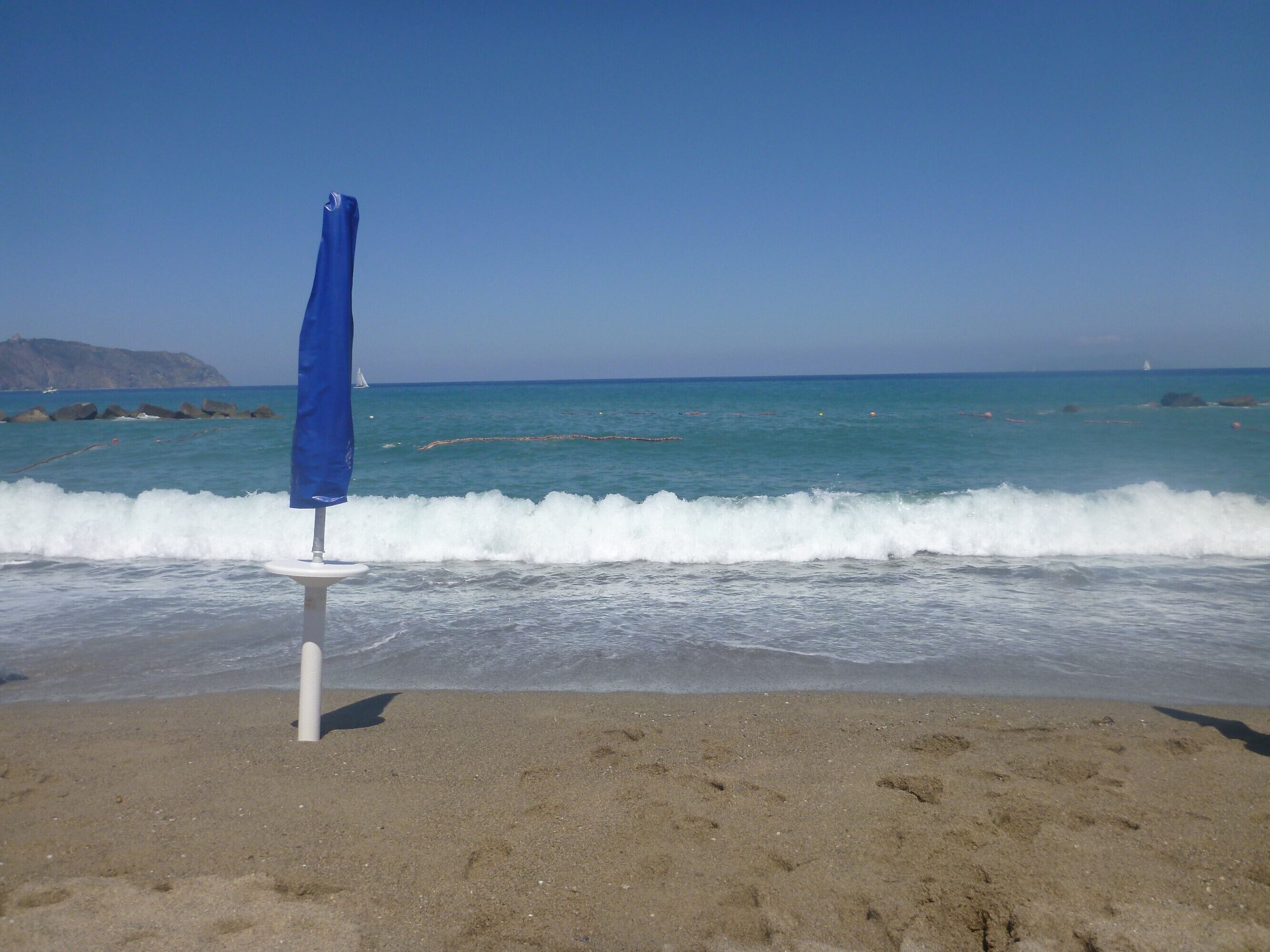 Una playa con un solo paraguas azul sobre un soporte blanco, olas rompiendo en la orilla y un cielo despejado.
