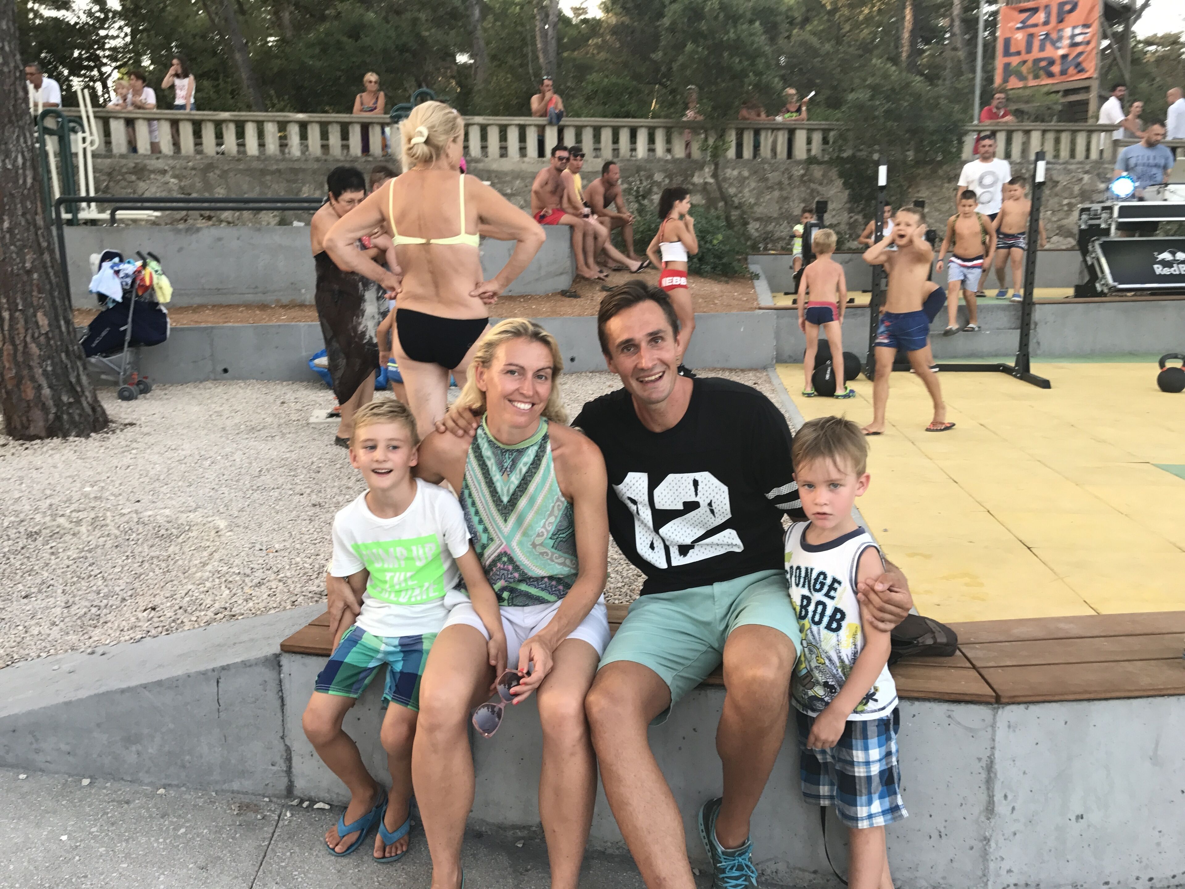 A family of four seated on a concrete bench in a public park with a stone wall and trees in the background.