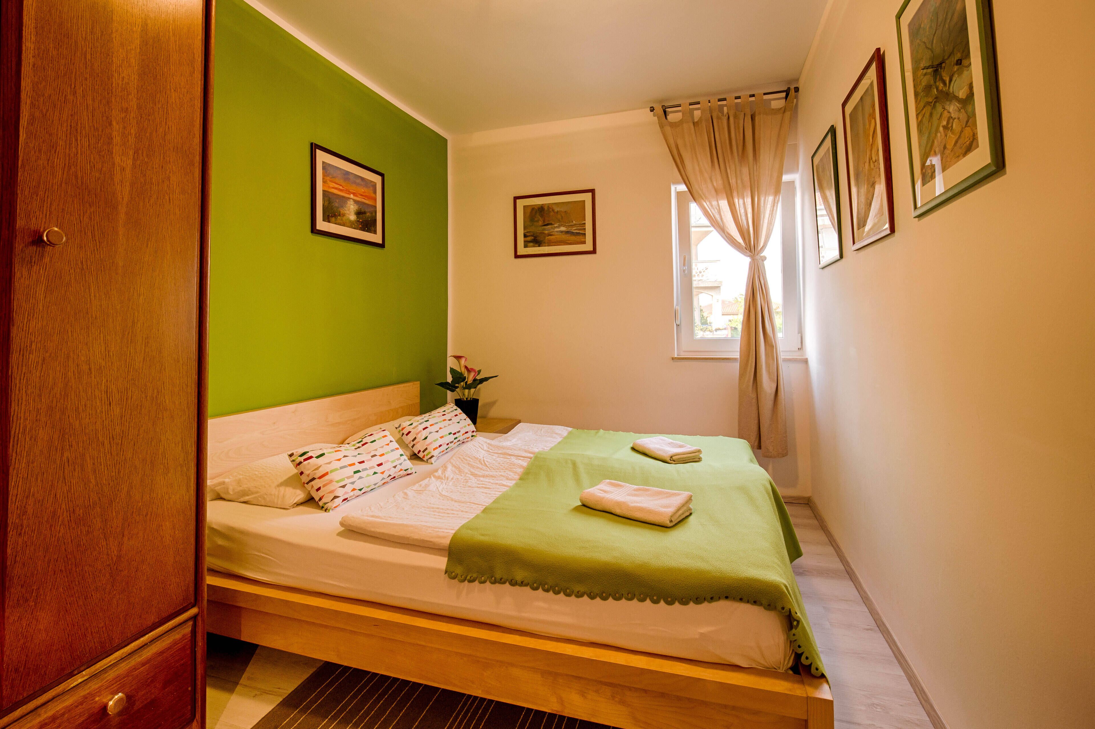 Een enkel bed met een groene deken, witte kussens en een kleine plant op het nachtkastje.