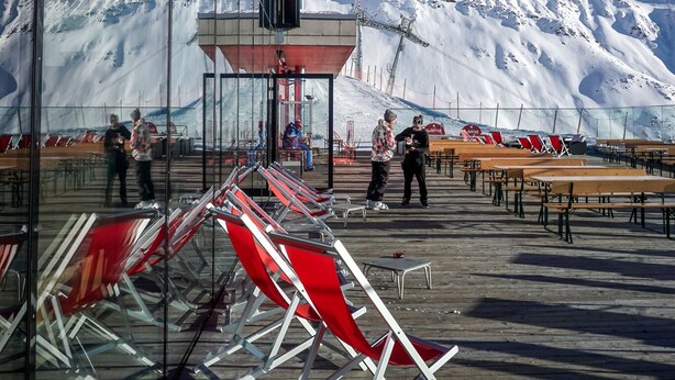 A snowy mountain backdrop with a glass-enclosed area featuring red chairs and tables, and a few people present.