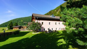 A large, two-story building with a sloped roof, surrounded by a well-maintained lawn and a forested hillside.