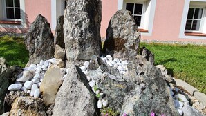 A cross on a rock with a solar panel, surrounded by stones and pink flowers, in front of a building with windows.