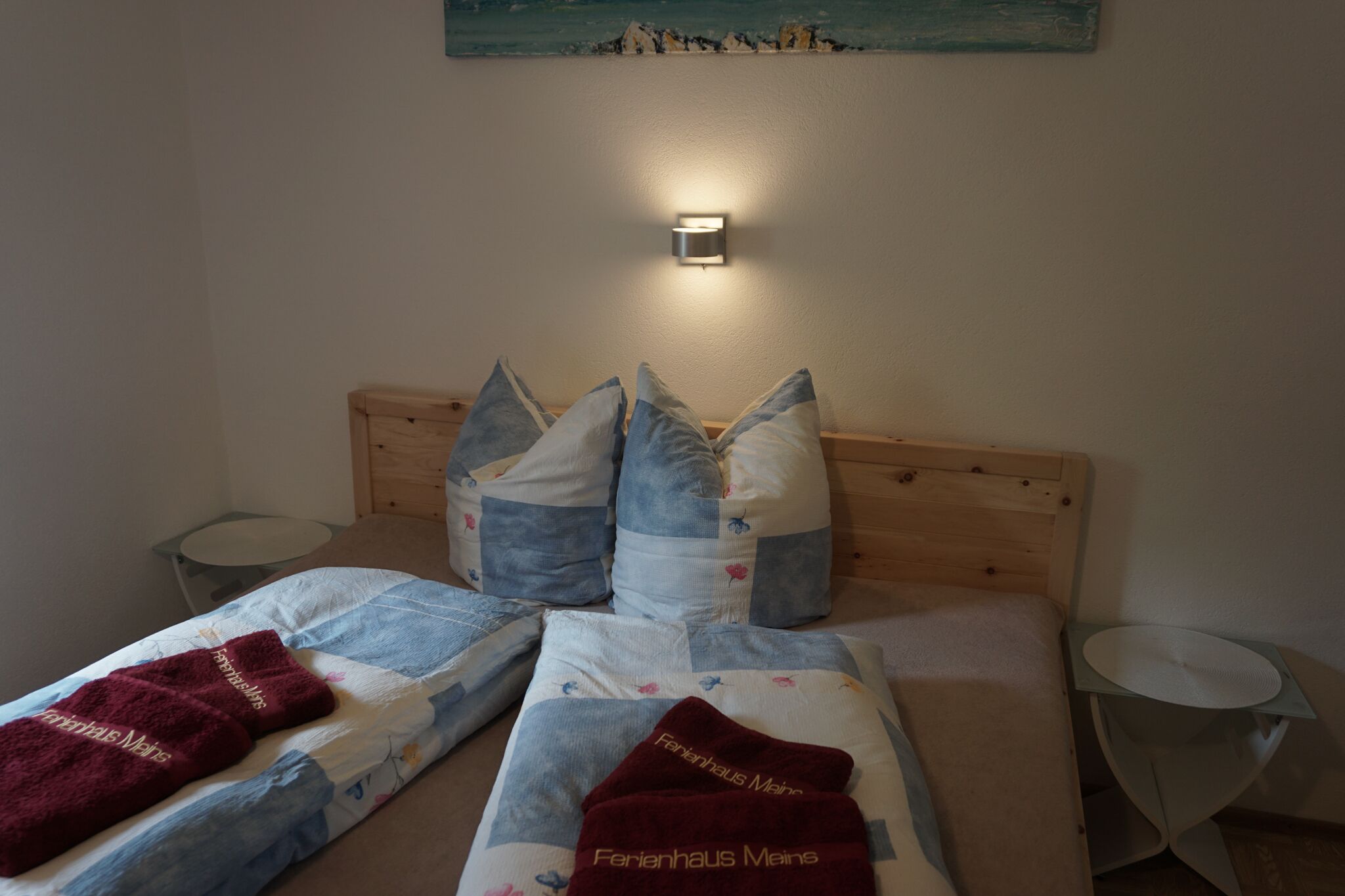 Ein ordentlich bezogenes Bett mit Holz Kopfteil, zwei Kissen und einer Bettdecke mit Aufdruck. Ein kleiner Nachttisch und eine Wandleuchte sind ebenfalls vorhanden.