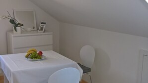 Une petite pièce moderne avec un plafond en pente, une table blanche sur laquelle est posée une assiette de fruits, une chaise blanche et un petit meuble blanc orné d’un tableau et d’une plante.