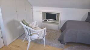 Une chambre à coucher comprenant un lit, une chaise en osier blanche, un bureau avec un écran d’ordinateur et une armoire.