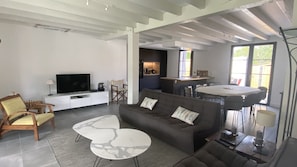 A modern living room with a sofa, coffee table, and TV. There is a kitchen area visible through an open door.