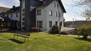 Un edificio de varios pisos con una torre angular destacada, un área de estar al aire libre cubierta y un patio ajardinado con un banco.