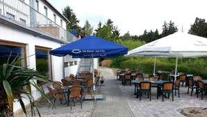 Un patio al aire libre con mesas y sillas, cubierto por sombrillas azules y blancas, y un edificio con una escalera.