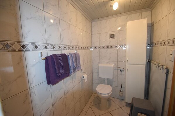 Um banheiro com azulejos de mármore branco, vaso sanitário, pia e um pequeno banquinho.