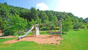 A playground with a slide, swings, and a climbing structure in a grassy area with trees in the background.