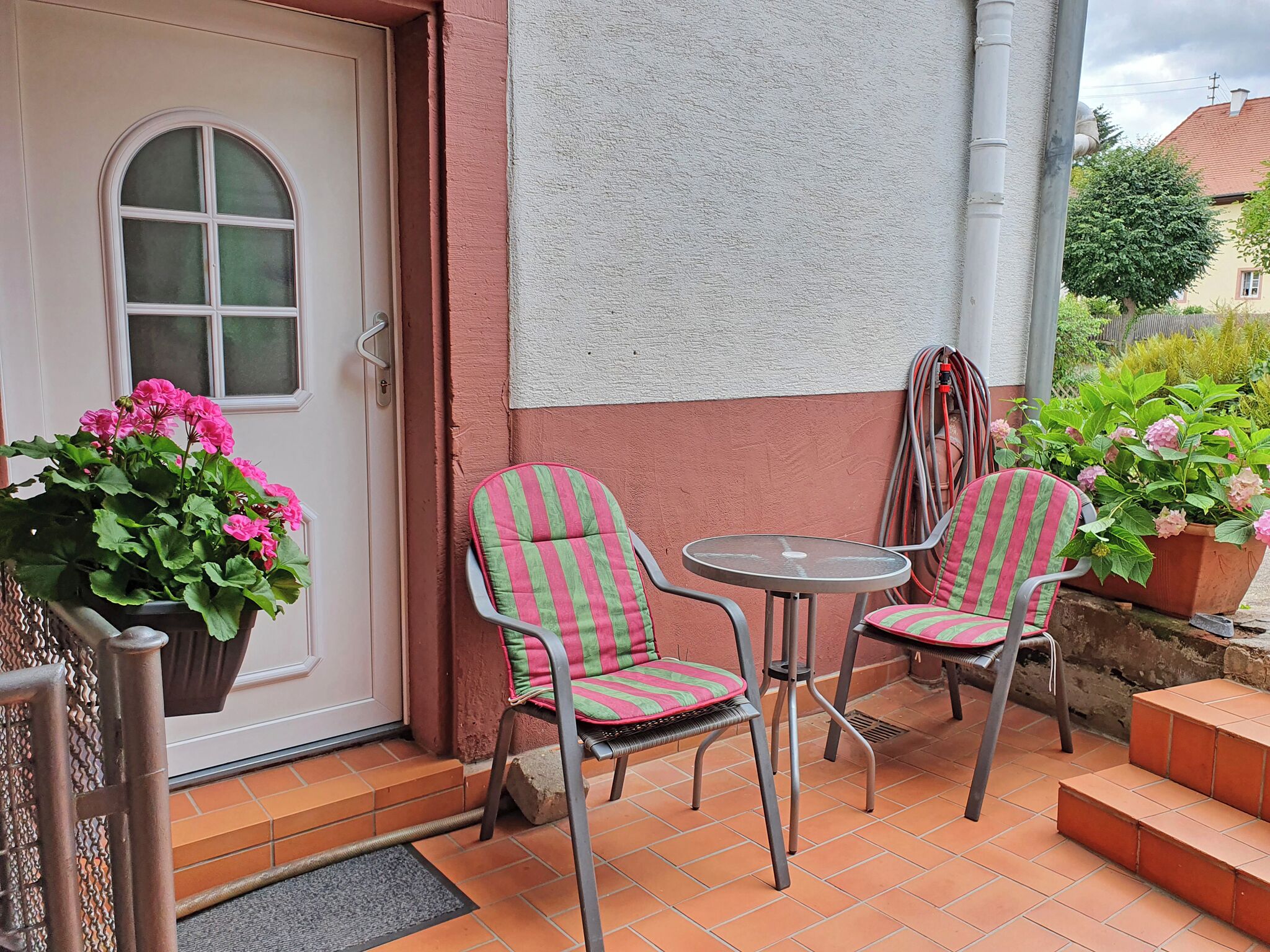 A small outdoor seating area with two striped chairs and a table, a potted plant with pink flowers, and a door with a window.