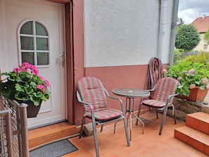 Un petit espace extérieur aménagé avec deux chaises à rayures et une table, un pot de fleurs orné de fleurs roses, et une porte dotée d’une fenêtre.