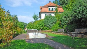 A house with a red roof, surrounded by lush greenery and a paved path leading to a small pool area.