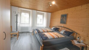 A bedroom with a wooden ceiling, two beds with patterned bedding, a coat rack, and a window with sheer curtains.