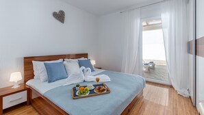 A neatly made bed with a tray of breakfast, a heart-shaped decoration on the wall, and a view of the beach through the window.