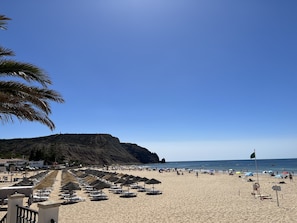 Ein Strand mit Reihen von Liegestühlen und Sonnenschirmen, einer Fahnenmast und einem Berg im Hintergrund.