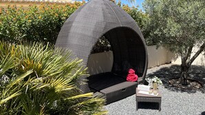 A wicker outdoor seating area with a cushioned bench and a small table, surrounded by gravel and plants. - Vila Marquês da Luz, ideal villa for guests looking for peace and quiet, sea view 1 floor (Luz)