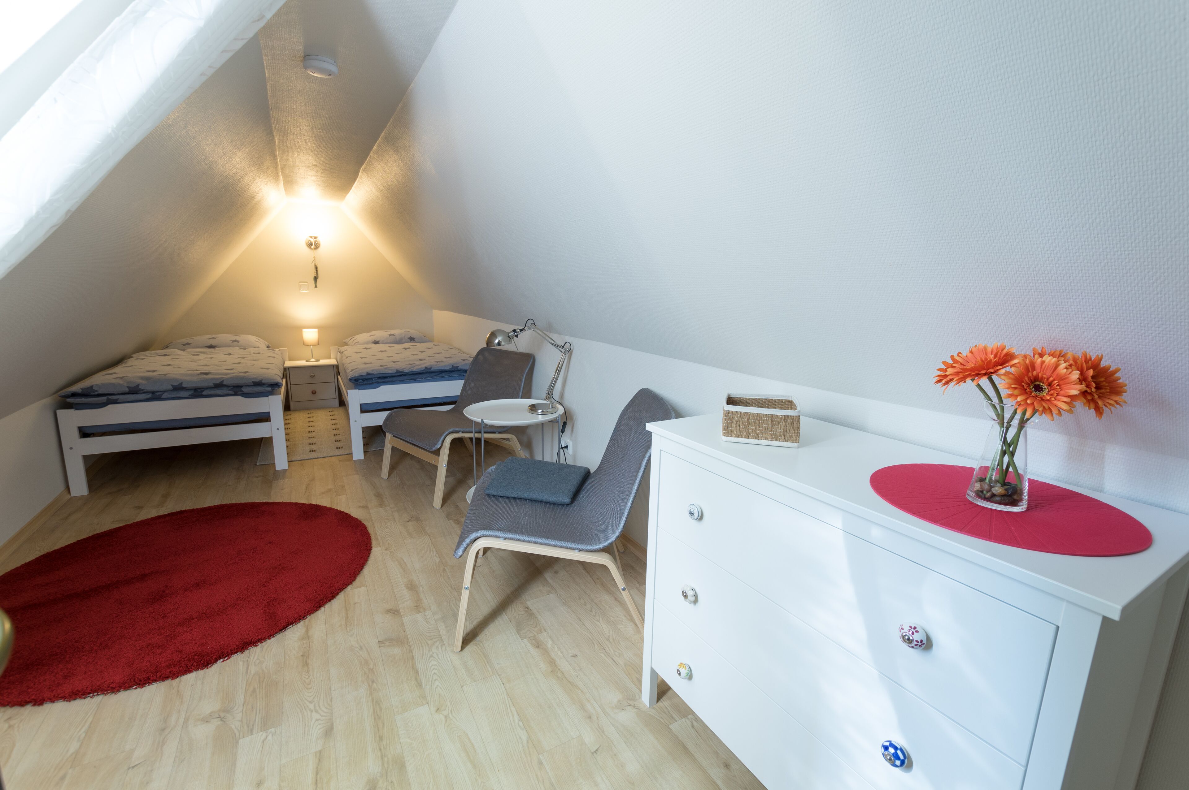 Une petite chambre mansardée, bien éclairée, avec un plafond en pente, deux lits, un bureau, une chaise, une commode et un tapis rouge.