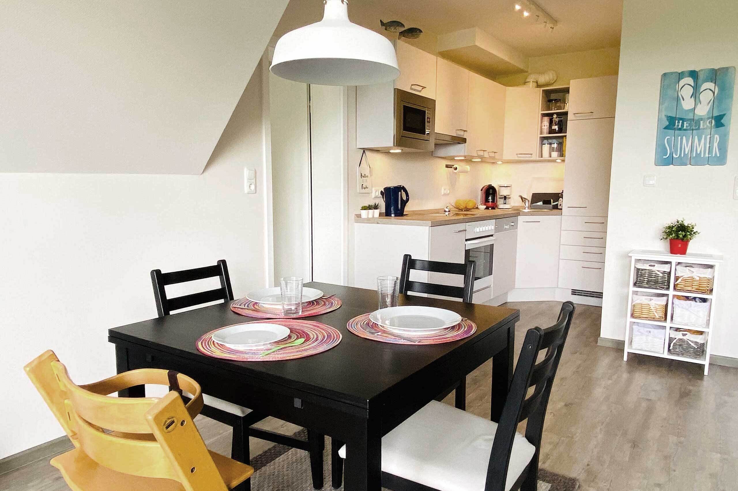Un comedor con una mesa y sillas negras, una lámpara colgante blanca y una cocina al fondo.