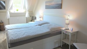 A bedroom with a bed, bedside lamps, a nightstand, a window with curtains, a radiator, a painting, and a television on the wall. - **** Apartment behind the dike (Westerdeichstrich)
