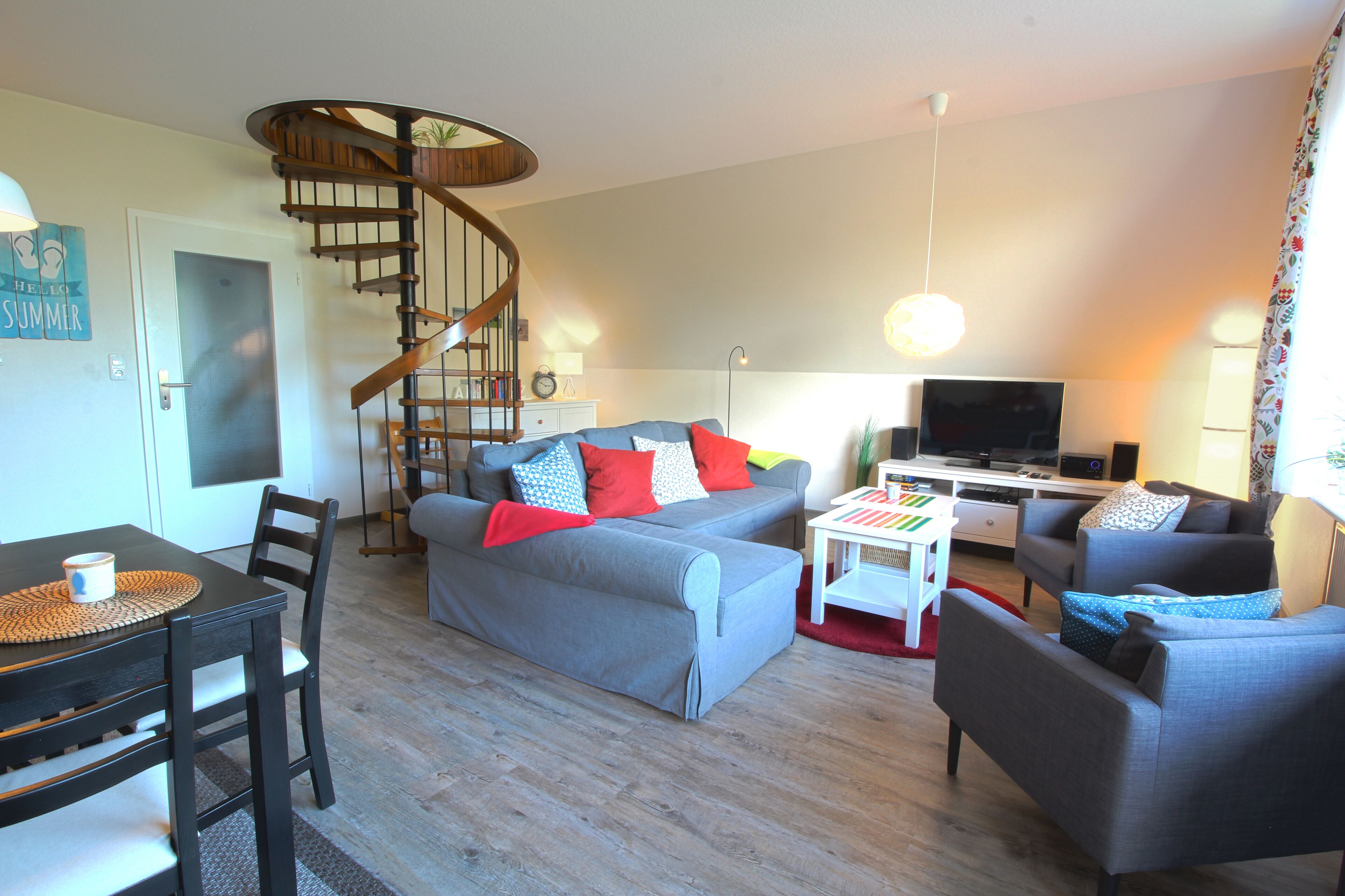 Ein Wohnzimmer mit Wendeltreppe, grauen Sofas, einem Fernseher und einem Essbereich mit Tisch und Stühlen.