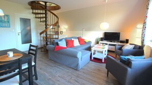 A living room with a spiral staircase, grey sofas, a TV, and a dining area with a table and chairs. - **** Apartment behind the dike (Westerdeichstrich)