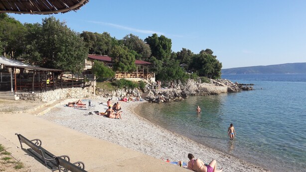 A pebbled beach with people sunbathing and swimming, a wooden structure with a red roof, and a rocky shoreline with trees. - Family-friendly country house Marie with heated pool, barbecue area, 5 persons (Krk)