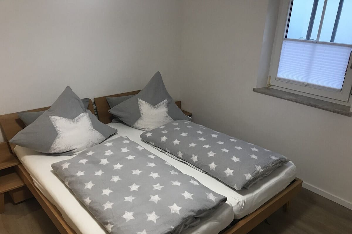 Ein Doppelbett mit einer grau-weißen Bettdecke und Kissen mit Sternenmuster, ein hölzernes Kopfteil und ein Fenster mit Jalousien.
