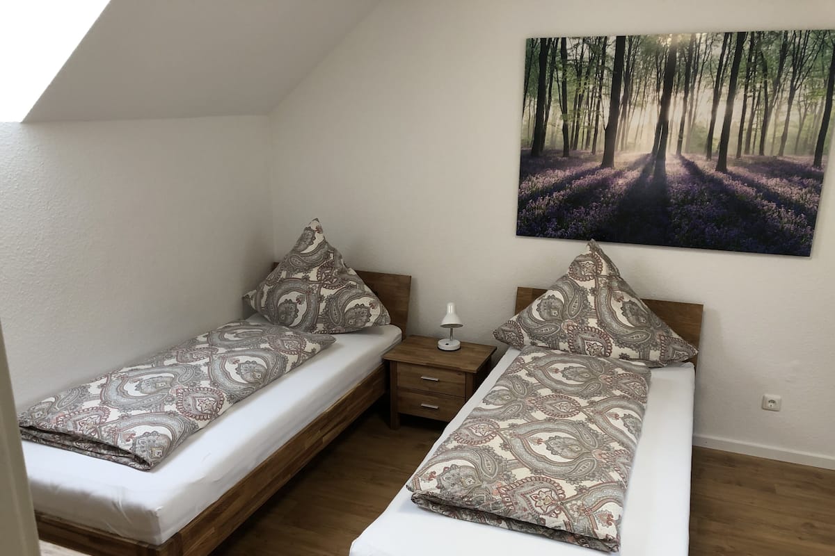 Ein Schlafzimmer mit zwei Einzelbetten, Holz-Kopfteilen, einem Nachttisch mit Lampe und einem Wandbild zum Thema Wald.