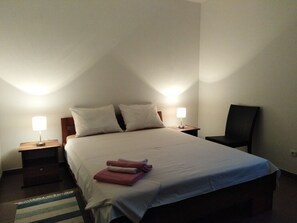 Ein ordentlich bezogenes Bett mit Kissen und gefalteten Handtüchern, zwei Nachttischlampen, ein Stuhl und ein Streifenmuster-Teppich in einem schlichten Raum mit weißen Wänden.
