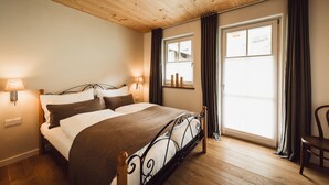 Ein Schlafzimmer mit Holzbalkendecke, einem Bett mit Metallgestell, einem Fenster mit Vorhängen und einer an der Wand befestigten Lampe.