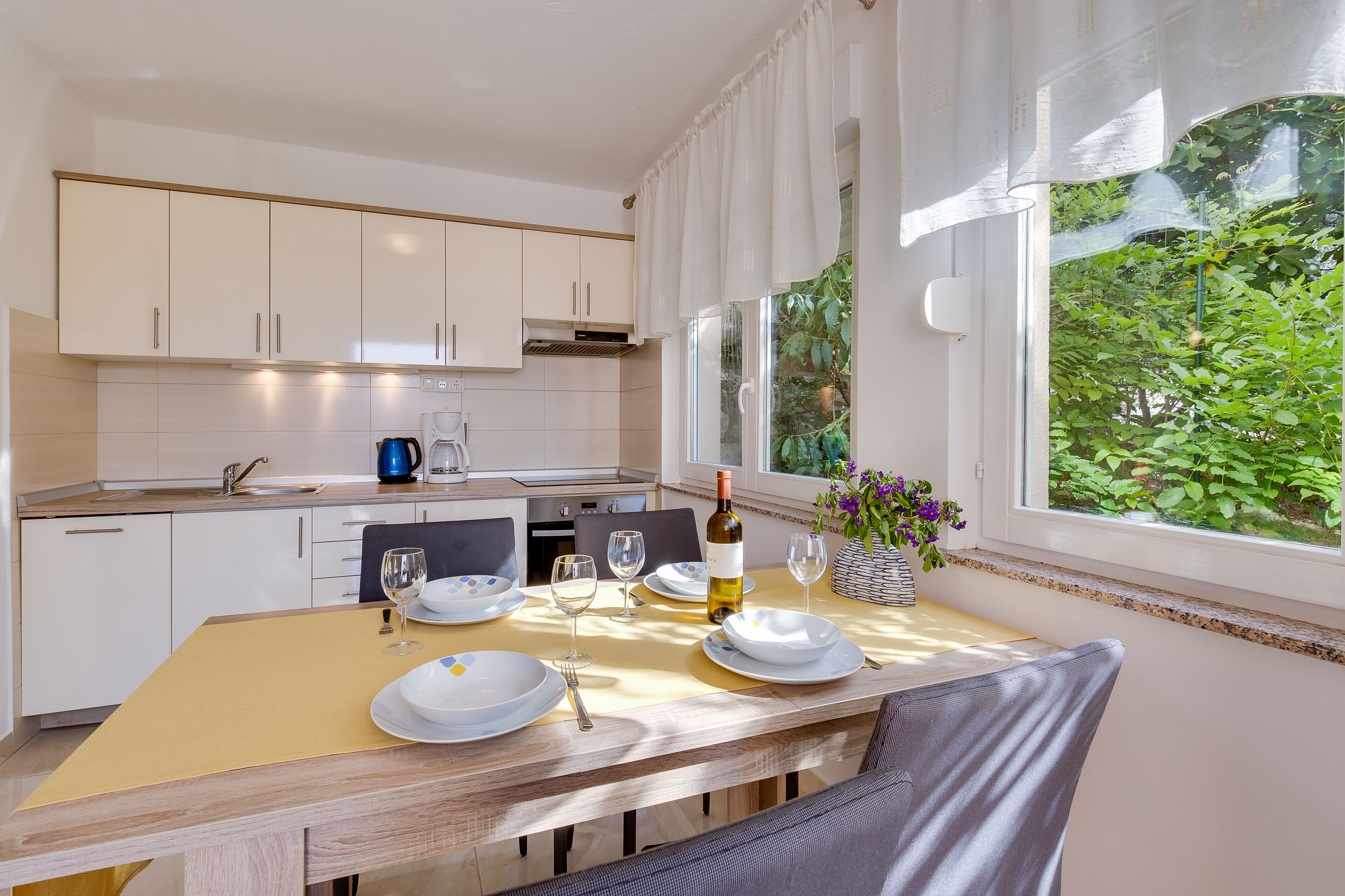 A modern kitchen with white cabinets, a dining table set for two, and a view of greenery through the window.