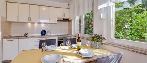 A modern kitchen with white cabinets, a dining table set for two, and a view of greenery through the window.