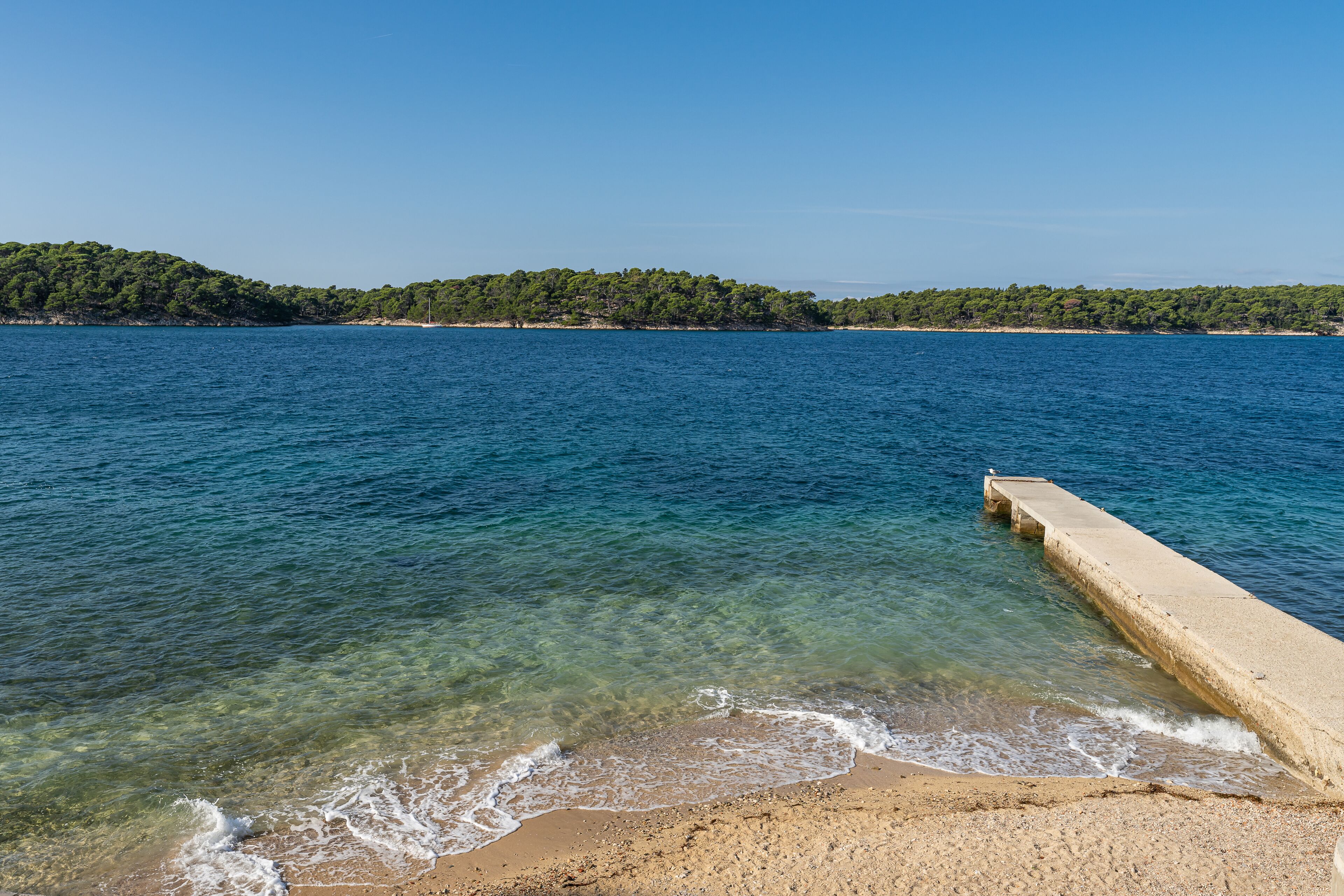 A clear view of a beach with calm waters, a concrete pier, and a forested island in the background.