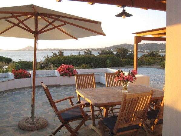 An outdoor patio with a table and chairs, a large umbrella, and a view of the sea and hills. - Naxos Beachvilla, a dreamhouse, 60m to sandy beach, big garden, top location (Naxos Town)