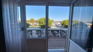 Une vue à travers une fenêtre, avec un ciel dégagé, quelques arbres et un parking aménagé avec des tables et des chaises.