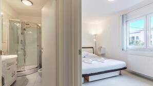 Une chambre d’hôtel moderne dotée d’une douche en verre, d’un lit blanc et d’une grande fenêtre laissant entrer la lumière naturelle.