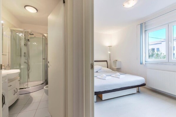 A modern hotel room with a glass-enclosed shower, a white bed, and a large window providing natural light.