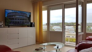 Un salon doté d’un grand téléviseur à écran plat, d’une table basse en verre, d’un canapé en cuir rouge et d’une porte-fenêtre coulissante donnant sur un balcon avec vue sur un port de plaisance.
